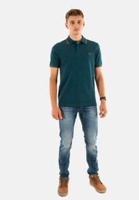 Jongeman staat rechtop en draagt een teal polo met gestreepte kraag, vervaagde blauwe jeans, bruine hoge schoenen en een zwarte horloge om zijn linkerpols.
