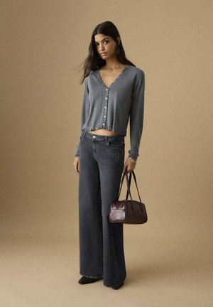 Cárdigan gris con escote festoneado y botones, combinado con vaqueros anchos oscuros con bordado floral. Bolso marrón que complementa el conjunto.