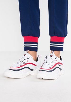 Osoba nosząca białe sneakersy FILA z czerwonymi i granatowymi akcentami, w połączeniu z granatowymi dresami z czerwono-białymi prążkowanymi mankietami.