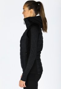 Gilet in velluto nero con colletto alto e cappuccio. Presenta tasche anteriori con zip e un design trapuntato. Indossato sopra un top scuro a maniche lunghe.