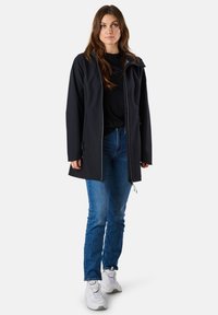 Czarna kurtka softshell z zamkiem błyskawicznym, dwoma bocznymi kieszeniami i kapturem, noszona na czarnej bluzce, w zestawie z niebieskimi dżinsami i białymi sneakersami.