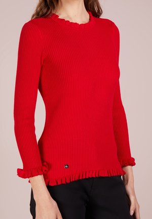 Kobieta ubrana w dopasowany czerwony prążkowany sweter z falbankami na kołnierzu, mankietach i dołu, w połączeniu z czarnymi spodniami.