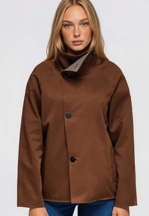 Une femme porte un manteau marron boutonné sur le devant et à col montant, avec un jean bleu, sur un fond uni et clair.