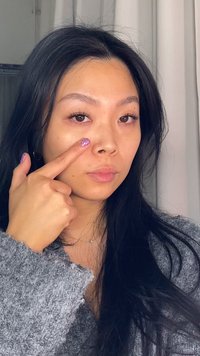 Femme aux longs cheveux noirs portant un pull gris, pointant sa joue avec une main ornée de vernis à ongles rose scintillant. Éclairage doux.