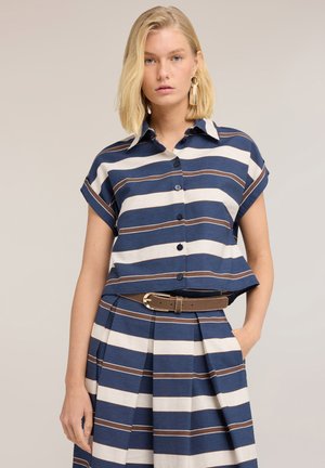 Femme blonde portant une chemise boutonnée à manches courtes rayée horizontalement bleu, blanc et marron et une jupe plissée assortie avec une ceinture marron.