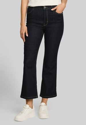 Personne portant un jean bootcut bleu foncé avec des coutures visibles et des baskets blanches à plateforme sur un fond gris uni.