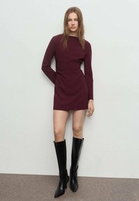 Vestido largo burdeos de manga larga con escote redondeado, cintura ajustada y detalles fruncidos. Combinado con botas de cuero negras hasta la rodilla. Textura suave.