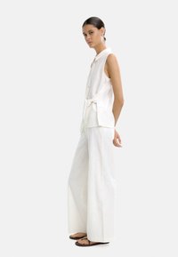Biała bezrękawnik z kołnierzykiem i guzikami z przodu, w połączeniu z szerokimi białymi spodniami. Teksturowana tkanina, luźny krój, prosty design.