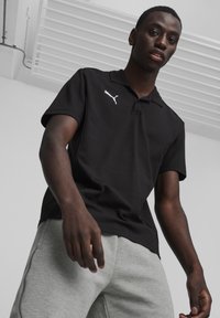 Zwarte polo met korte mouwen, voorzien van een wit Puma-logo op de linkerkant van de borst, gecombineerd met grijze joggingbroeken. Eenvoudig ontwerp, gemaakt van katoen.