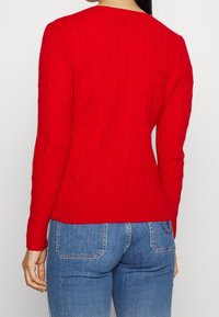 Pull rouge en tricot avec un motif torsadé, poignets et ourlet côtelés. Porté avec un jean en denim bleu, mettant en valeur une silhouette ajustée vue de dos.