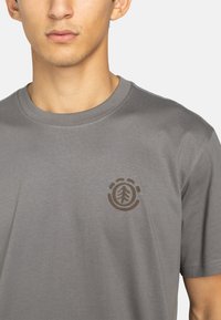 Grijze katoenen t-shirt met een ronde hals en korte mouwen, voorzien van een bruine boomlogo op de linkerkant van de borst. Gladde textuur.