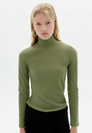 Olive green long-sleeve turtleneck, ribbed texture, fitted design, soft fabric, worn with a black bottom, model with earrings, neutral background.
