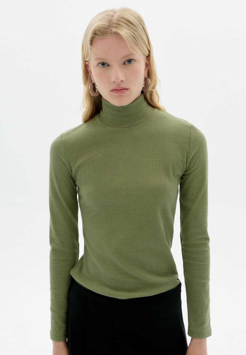 Olivgrüner langärmeliger Rollkragenpullover, gerippte Textur, figurnaher Schnitt, weicher Stoff, mit einem schwarzen Unterteil getragen, Modell mit Ohrringen, neutraler Hintergrund.