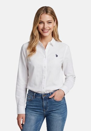 Lächelnde Frau mit blondem Haar, die ein weißes Hemd mit Knöpfen und Logo sowie blaue Jeans trägt, Hand in der Jeanstasche, vor einfarbigem Hintergrund.