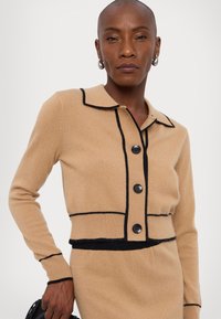 Beige strikket cardigan med svart trim, med krage, knappfront og lange ermer. Glatt tekstur med kontrasterende aksenter.
