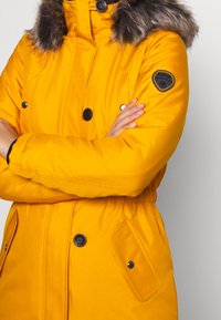 Manteau d'hiver jaune vif avec boutons noirs, capuche doublée de fourrure, poches à rabats avec pressions, et écusson de marque sur le haut du bras.