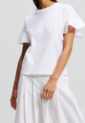 Valkoinen lyhythihainen puuvillainen t-paita, jossa on röyhelöhihat ja pyöreä pääntie. Etupuolella on hillitty kohokuvioitu logodetaili.