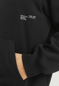 Sweatshirt noir avec une poche avant, des poignets côtelés et un design de texte blanc avec des coordonnées géographiques et des étiquettes. Texture de tissu lisse.