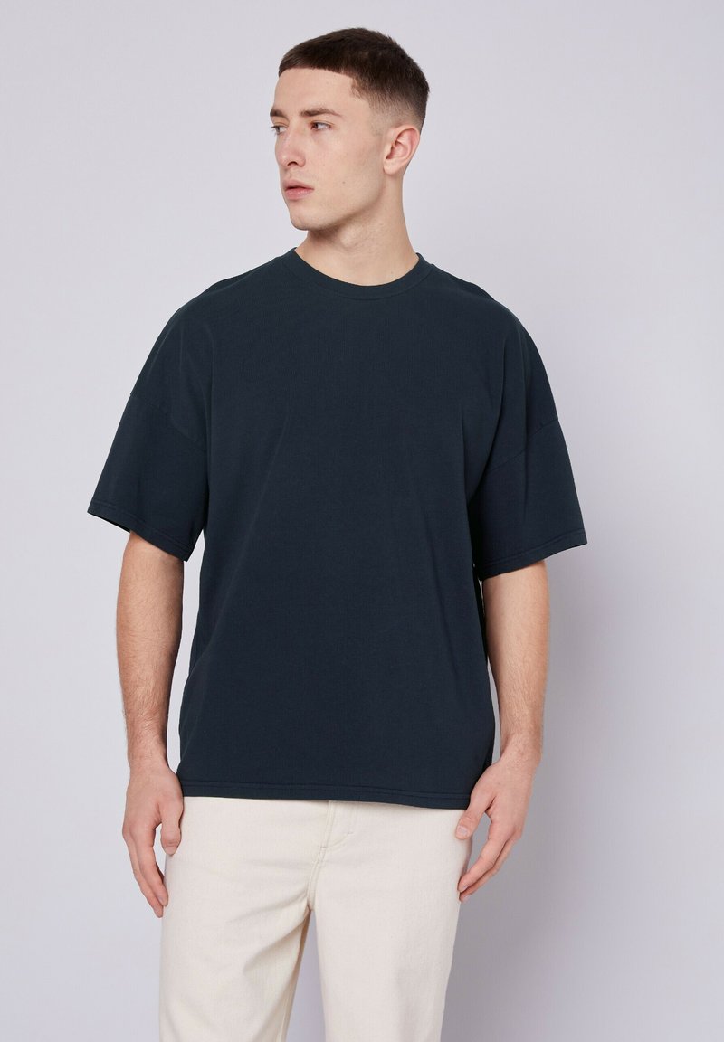 Juodas per didelis marškinėlis, tekstūruotas medvilnės audinys, trumpomis rankovėmis, apvalia iškirpte, laisvas kirpimas. Modelis vilki marškinėlį su šviesios spalvos kelnėmis.