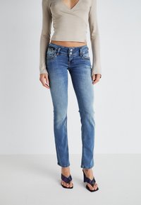 Jean en denim bleu avec une finition délavée, design �à cinq poches et coutures contrastées. Associé à un haut à manches longues beige.