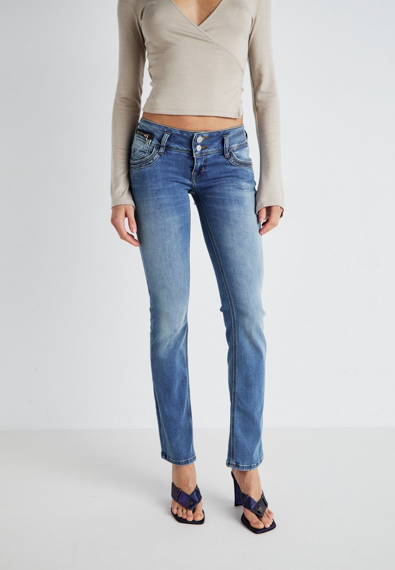 Jean en denim bleu avec une finition délavée, design à cinq poches et coutures contrastées. Associé à un haut à manches longues beige.
