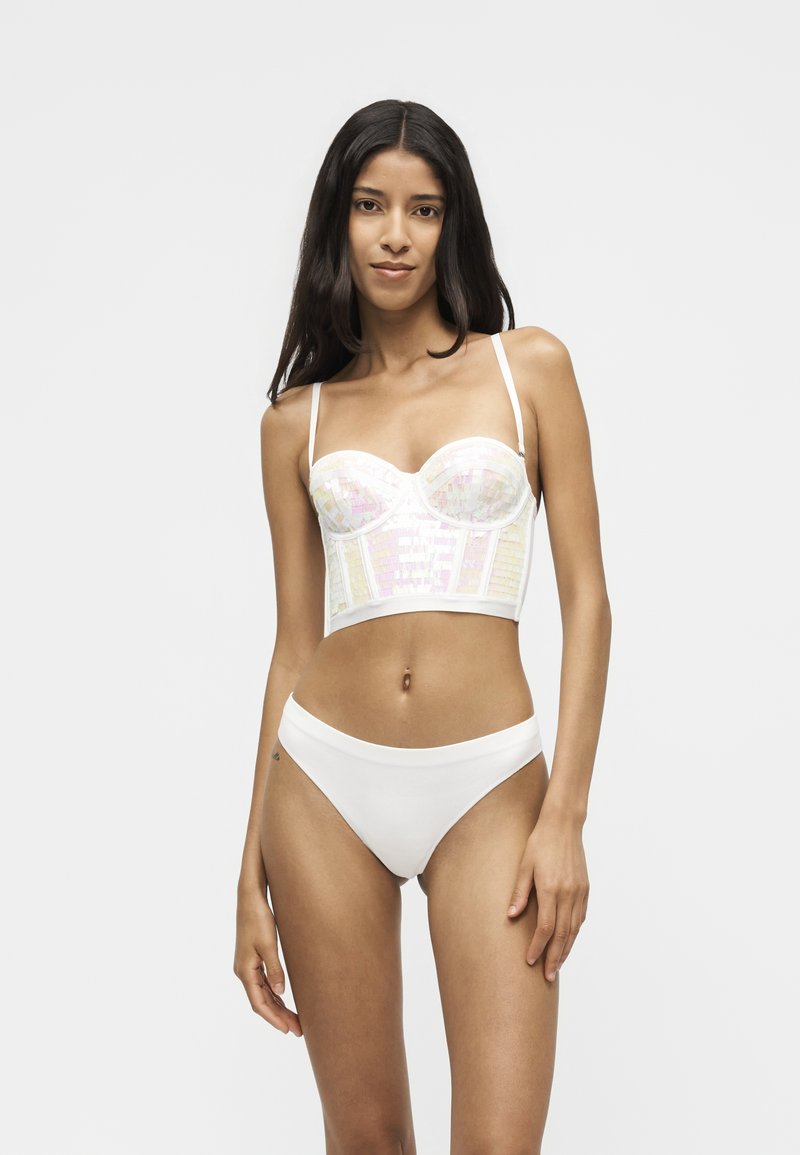 Femme aux longs cheveux foncés portant un bustier blanc irisé et un sous-vêtement blanc simple style bikini sur un fond clair uni.