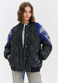 Prešívaná čierna puffer bunda s modrými akcentami a vzorovanými detailmi na pleciach. Obsahuje patentky, rebrované manžety a dekoratívny zips s strapcom.