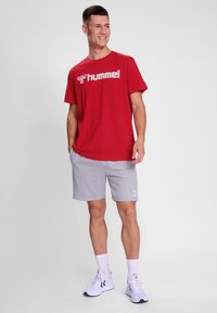 Rød kortærmet t-shirt med hvid "hummel" logo, parret med grå shorts og hvide sportsko. Enkle teksturer, afslappet pasform.