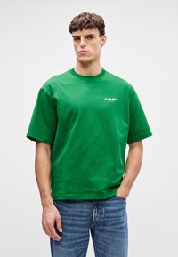 T-shirt verde de algodão com gola redonda, mangas curtas e corte solto. Apresenta bordado de logótipo branco na parte frontal. Combinado com jeans de ganga azuis.