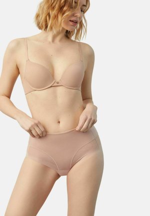 Mujer ajustándose unas braguitas de talle alto de tono nude, con un sujetador a juego con aro, con cabello rubio hasta los hombros, sobre un fondo liso.