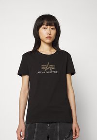 T-shirt en coton noir avec le logo "ALPHA INDUSTRIES" en or métallisé et un design géométrique sur le devant. Manches courtes, col rond.