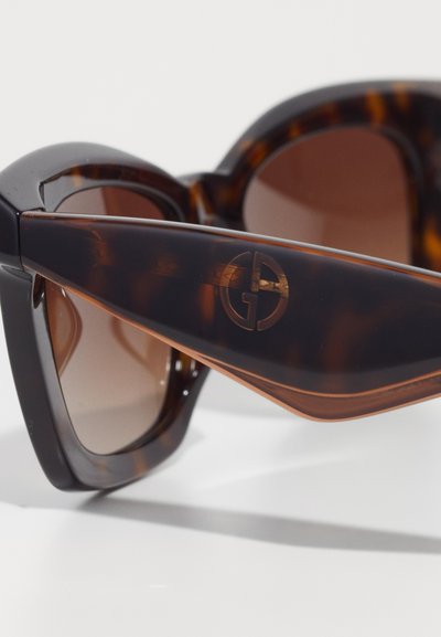 Lunettes de soleil en écaille de tortue de forme œil de chat avec des verres dégradés bruns, présentant un logo circulaire en relief sur la branche.
