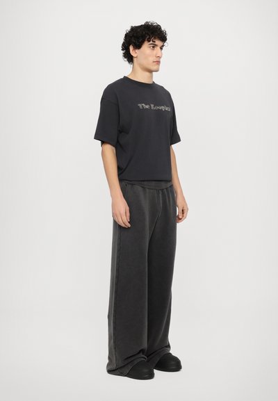 Sort kortærmet t-shirt med logo, parret med brede mørkegrå bukser og sorte slip-on sko. Glat stof, afslappet pasform, minimalistisk design.