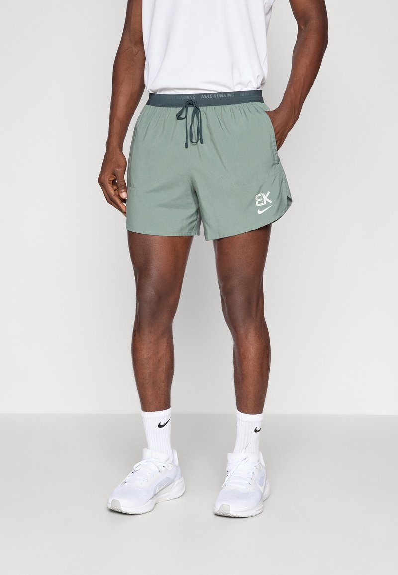 Homme portant un short de course Nike vert sauge, des chaussettes blanches Nike et des chaussures de sport blanches, debout avec une main dans la poche.