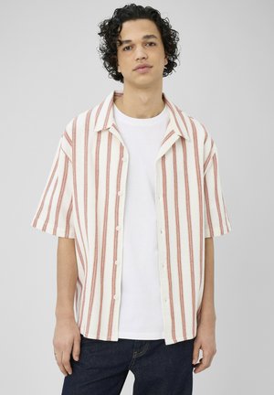 Jeune homme aux cheveux bouclés et foncés, portant un t-shirt blanc sous une chemise blanche à manches courtes avec des rayures rouges verticales et un jean foncé.