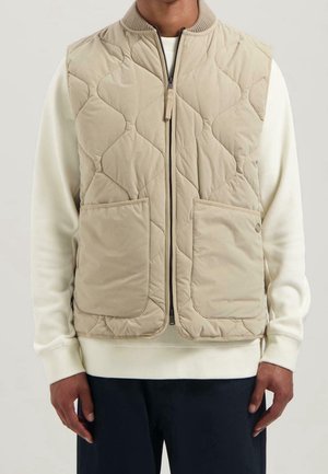 Uomo che indossa un gilet trapuntato beige con zip sopra una camicia bianca a maniche lunghe e pantaloni scuri, in piedi davanti a uno sfondo neutro.