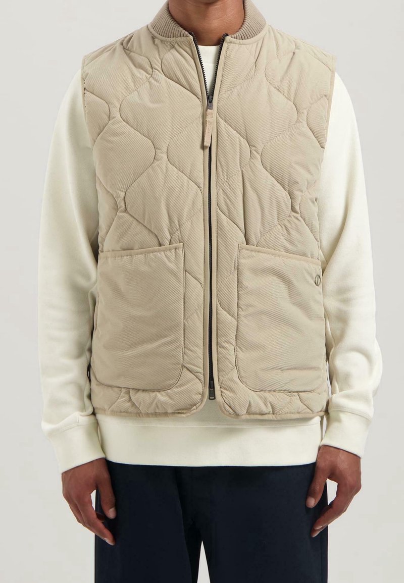 Homme portant un gilet matelassé beige zippé par-dessus une chemise blanche à manches longues et un pantalon foncé, debout devant un fond neutre.