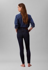 Jeans skinny din denim închis, cu talie înaltă, asortați cu o cămașă din denim albastru, cu mâneci suflecate și guler cu nasturi, văzuți din spate.
