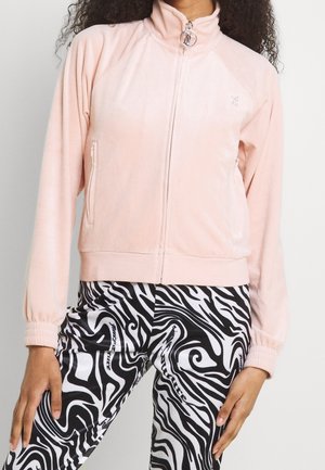 Femme portant une veste zippée rose clair et des leggings noirs et blancs à motifs abstraits en spirale, debout les mains détendues le long du corps.