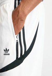 Bílé sportovní šortky s třemi černými pruhy po stranách, kapsou na zip a vyšitým logem. Hladká tkanina a elastický pás.
