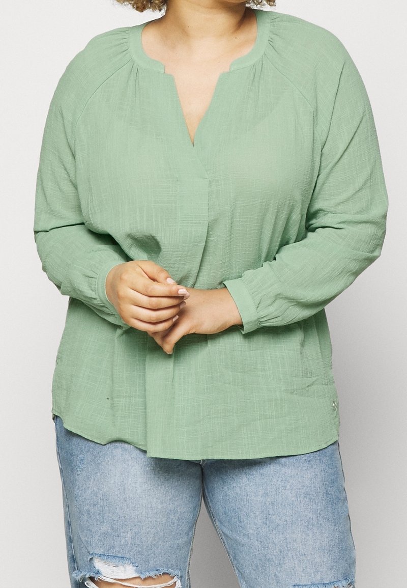 Blusa verde chiaro a maniche lunghe con scollatura a V e collare arricciato. Presenta un tessuto testurizzato e un orlo arrotondato, abbinata a pantaloni corti in denim strappati.