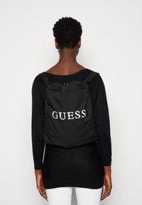 Černý batoh z nylonu s logem "GUESS" ve stříbrné barvě, vybavený nastavitelnými popruhy a horním zipem. Minimalistický design.