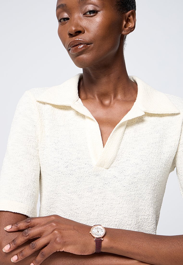 Reloj de pulsera con una esfera plateada, detalles en dorado y una correa trenzada en color marrón. El modelo lleva una camiseta polo texturizada de color crema claro.