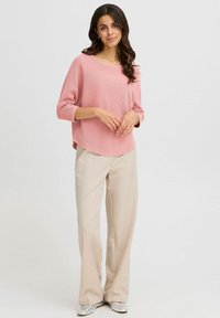 Maglione rosa chiaro, texture a maglia con scollatura ampia e maniche a tre quarti, abbinato a pantaloni beige chiari a taglio morbido e scarpe argentate.
