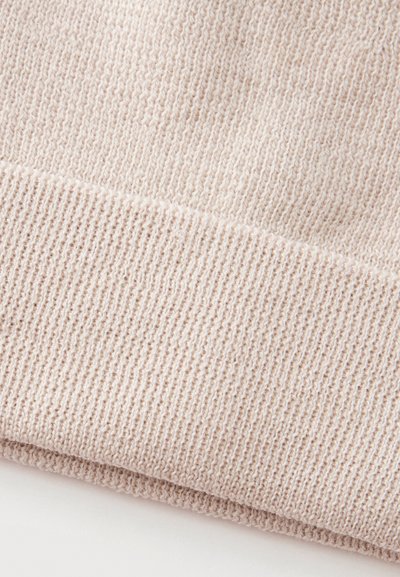 Gros plan sur un tissu tricoté côtelé beige doux avec des coutures visibles et une texture, montrant un détail d’ourlet plié.