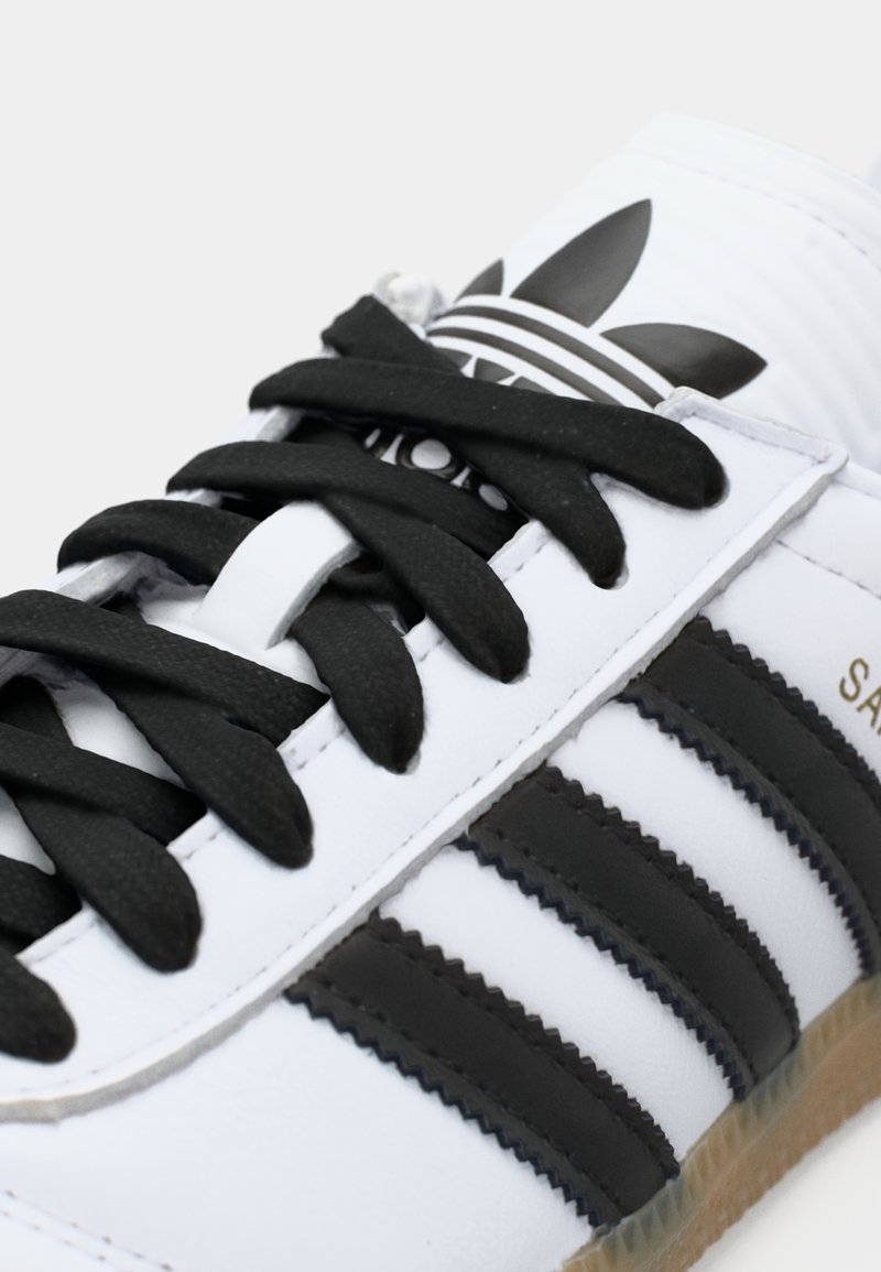 Weißer Sportschuh mit schwarzen Streifen und Schnürsenkeln. Verfügt über ein strukturiertes Obermaterial, Logo-Akzente und eine Gummisohle.