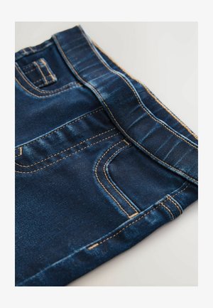 Sötétkék farmernadrág narancssárga varrással, amely derékpánttal, elülső zsebbel és sima textúrával rendelkezik. Közelkép a szövet részleteiről.