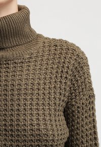 Jersey de cuello alto de punto en verde oliva, con un patrón texturizado y un cuello acanalado. La tela parece cálida y gruesa.