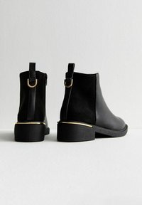 Botas de tobillo de cuero negro con un acabado suave, detalles en dorado en el tacón y una cremallera en el costado. Material superior ligeramente texturizado.