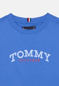 Μπλε βαμβακερό T-shirt με στρογγυλή λαιμόκοψη. Στο μπροστινό μέρος εμφανίζεται το «TOMMY» σε άσπρο και το «HILFIGER» σε κόκκινο, με ετικέτα λογότυπου ορατή μέσα στον γιακά.